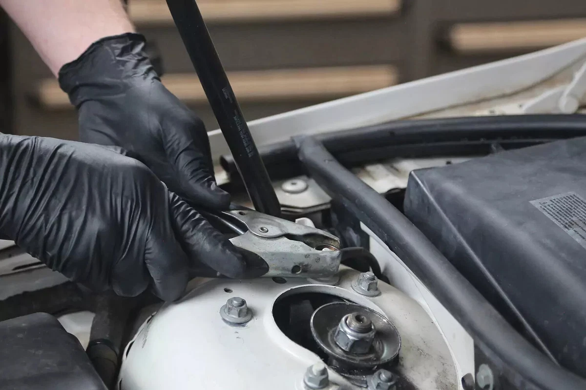 A mechanic wearing black gloves disconnects a black handled jumper cable from a grounding point on a car.