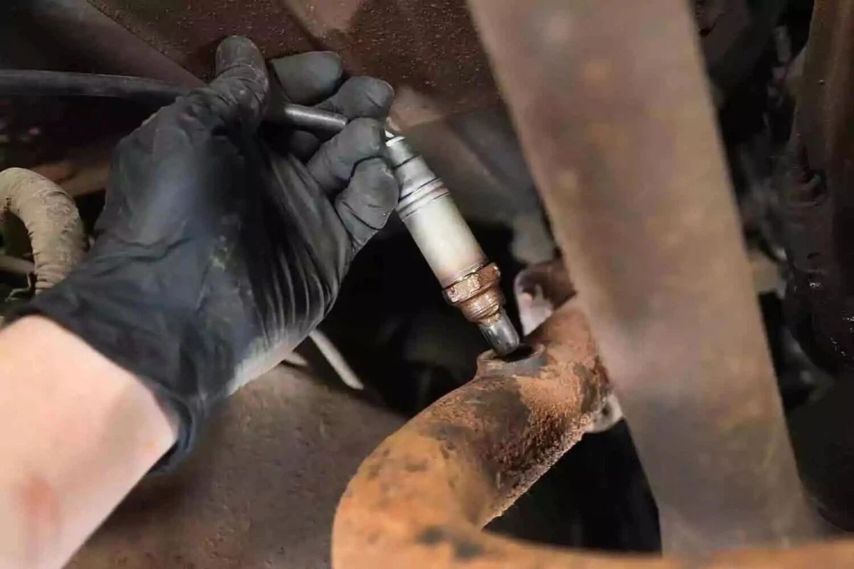 A mechanic wearing a black nitrile glove inserts the new oxygen sensor, its threads coated with anti-seize compound, into the exhaust pipe bung.
