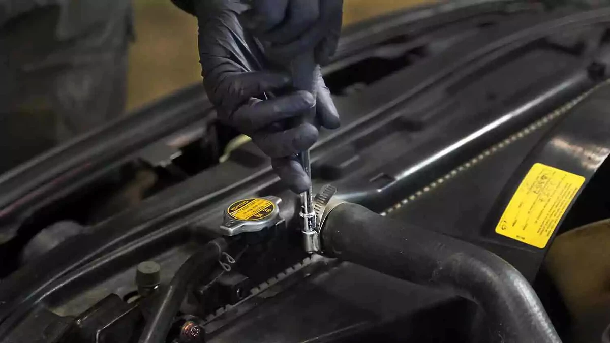 A mechanic wearing nitrile gloves reattaches an upper radiator hose to a car radiator with a socket wrench.