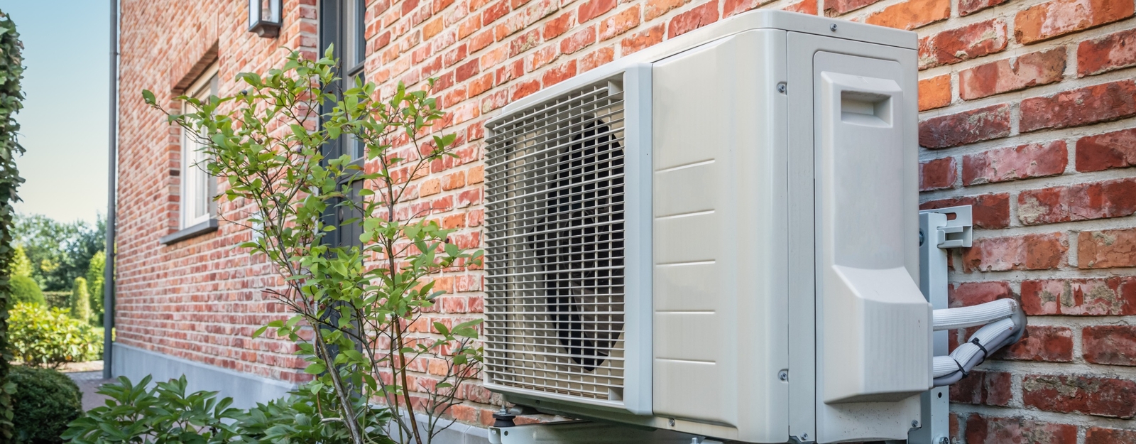 Außenansicht einer Wärmepumpe an der Ziegelaußenwand eines Hauses, umgeben von grünen Pflanzen und einem gepflegten Garten