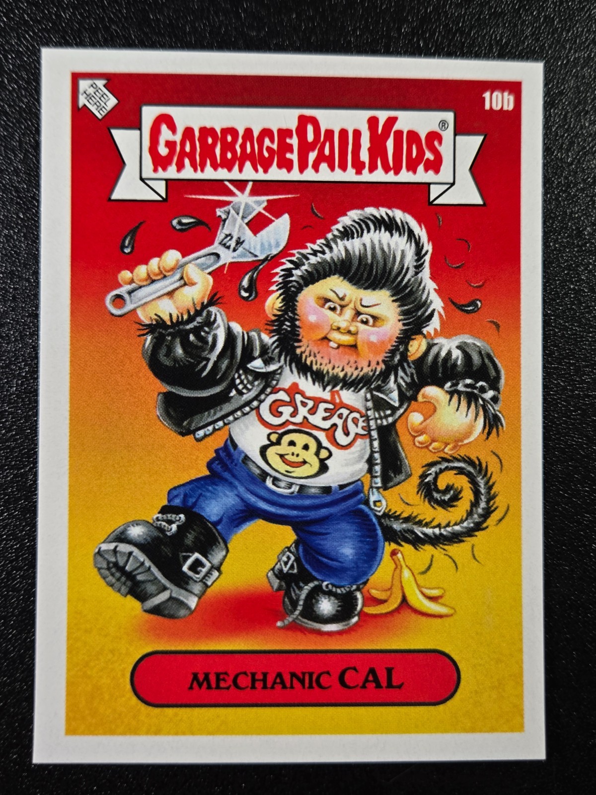 Greaser Greg comic card from Garbage Pail Kids, blue shirt, black hair, holding wrench, yellow-red circle background.