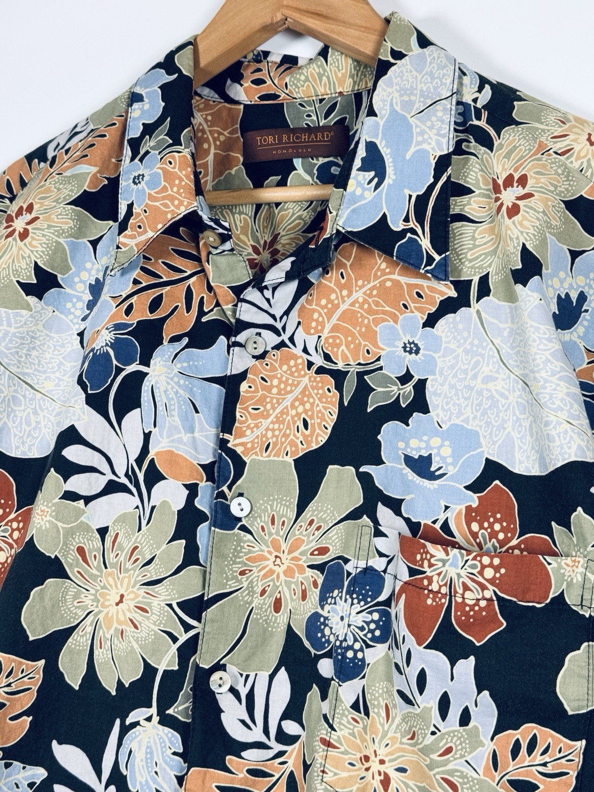 Black Hawaiian shirt with orange, blue, and green floral print, front pocket, and “Tori Richard” label on hanger.