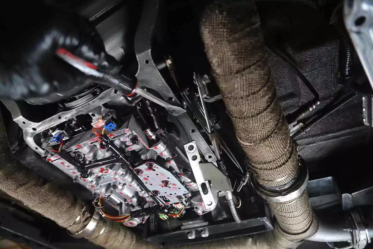 A mechanic wearing a black glove uses a gasket scraper to clean away pieces of the old transmission filter.