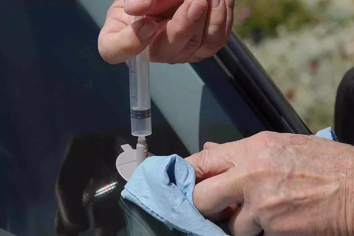 A vehicle-owner uses a small windshield chip repair device to fill a chip with resin, while using a blue disposable shop towel to contain overflow.