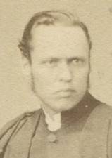 MAN IN GOWN AT TABLE WITH CAP, BOOK. ST BEES COLLEGE, ENGLAND.  