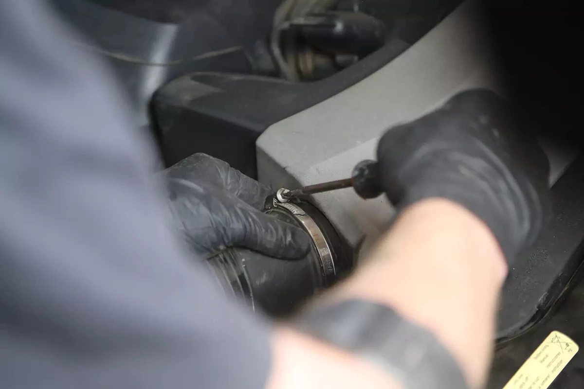 A mechanic wearing a black glove uses a screwdriver to tighten the hose clamp around the air tubing leading to the air filter box.