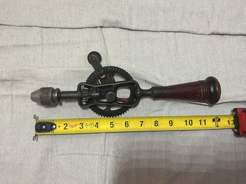 Vintage black and red hand crank drill with worn wooden storage handle, yellow tape measure, and visible branding.