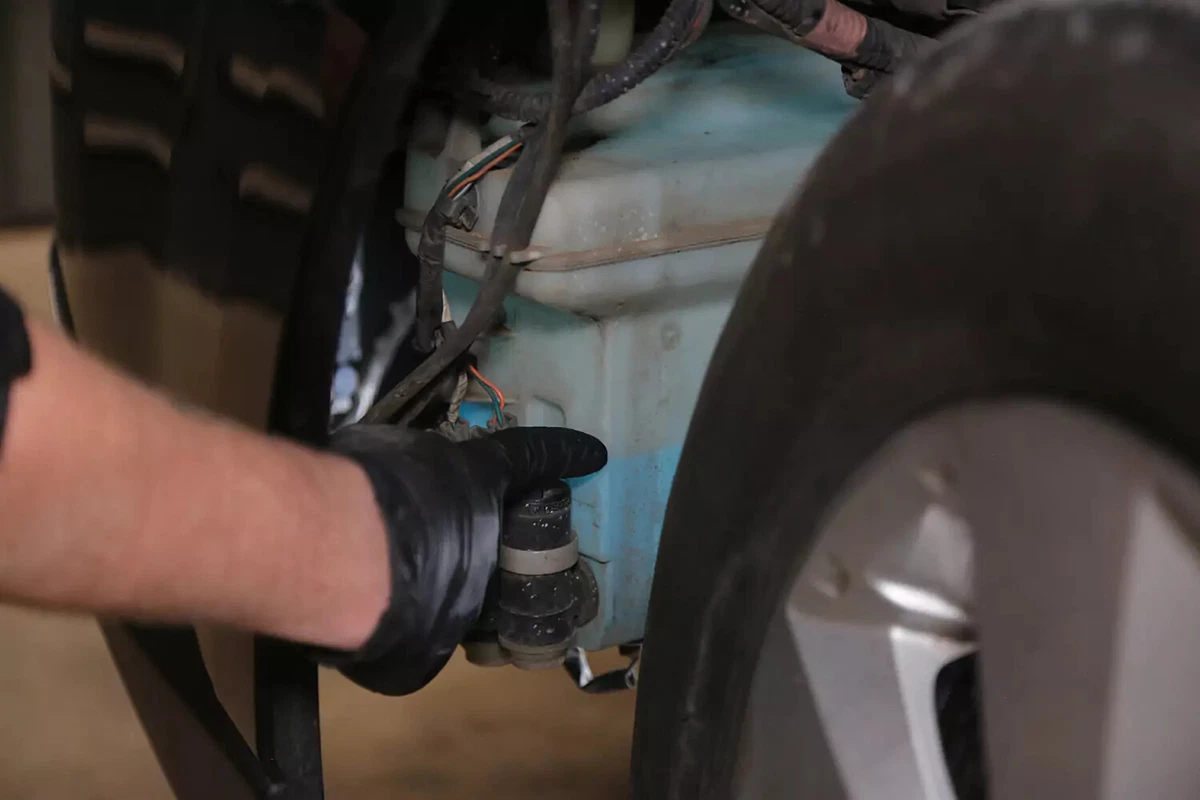 A mechanic wearing a black glove pointing to a vehicle's windshield fluid reservoir.