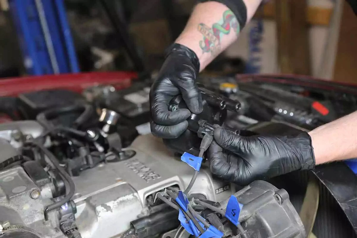 A mechanic wearing black nitrile gloves removing the spark plug wires from the distributor cap. The spark plug wires are labeled with blue tape.