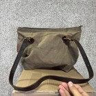 Brown canvas crossbody bag with black leather strap, front zipper pocket, and two brass rings, on a wooden base.