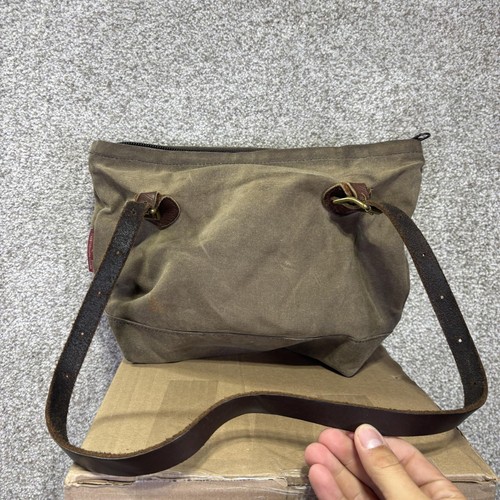 Brown canvas crossbody bag with black leather strap, front zipper pocket, and two brass rings, on a wooden base.