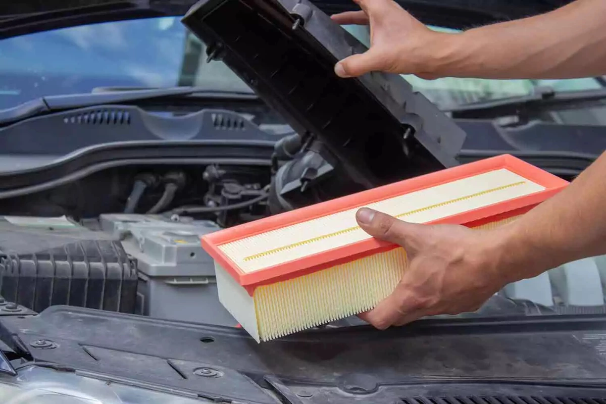 A mechanic installing a new engine air filter. He is opening the air cleaner box with his right hand while holding the filter in his left hand.
