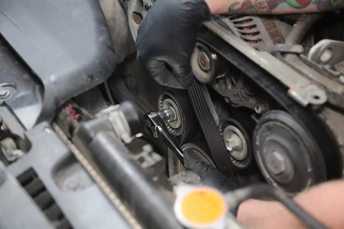 A mechanic wearing a black glove uses a ratchet to loosen the engine's serpentine belt tensioner.