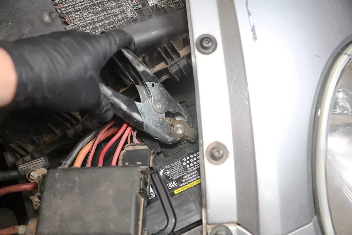 A mechanic wearing a black glove attaches a black handled jumper cable to the negative charging post of a car battery.