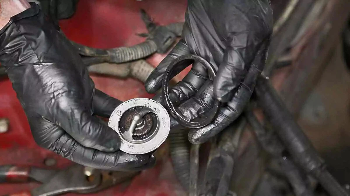 A mechanic wearing black nitrile gloves holding a new thermostat and gasket.
