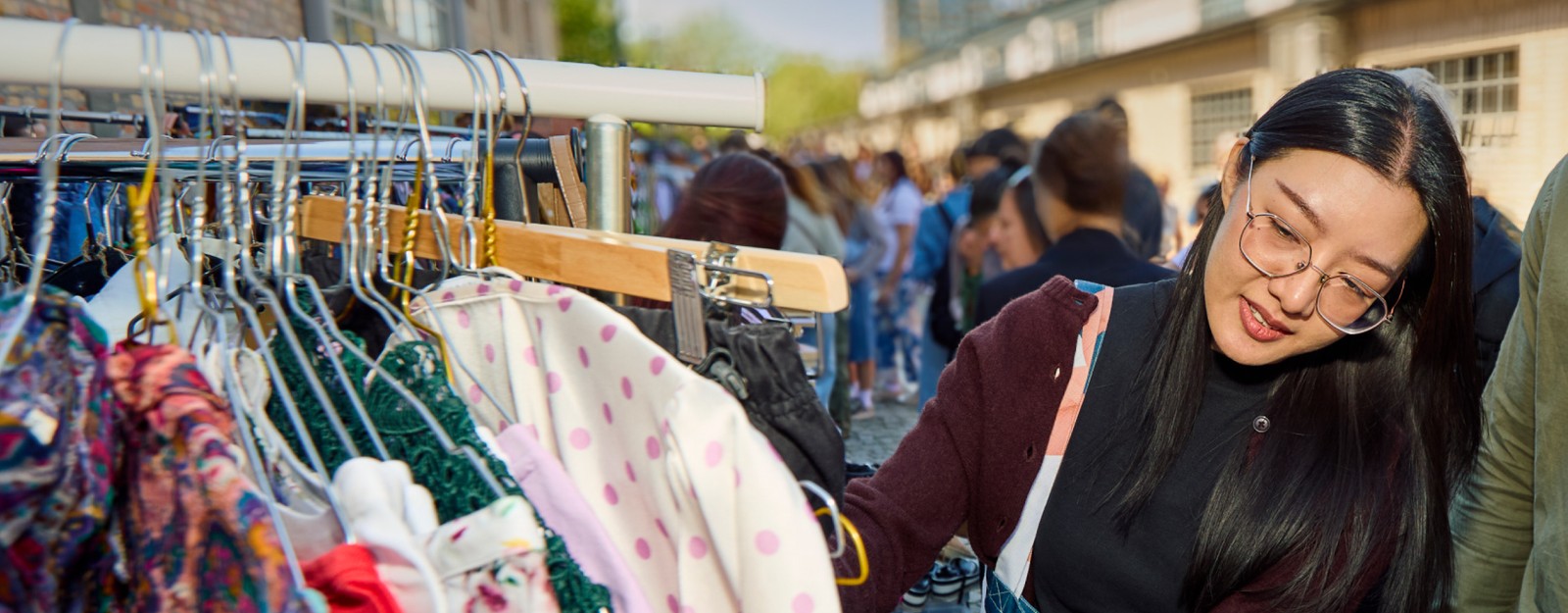 Eine junge Frau mit Brille stöbert lächelnd an einer Kleiderstange auf einem gut besuchten Outdoor-Flohmarkt nach Secondhand-Kleidung.