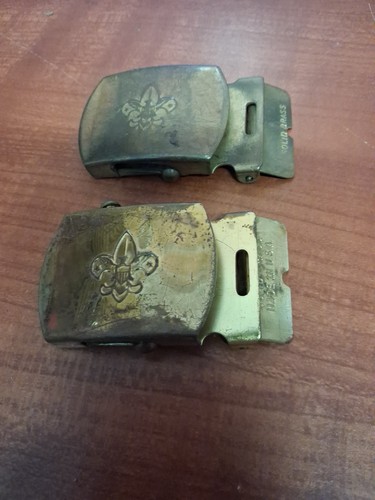 Two worn brass BSA Boy Scouts belt buckles with embossed fleur-de-lis logo on a wooden surface.