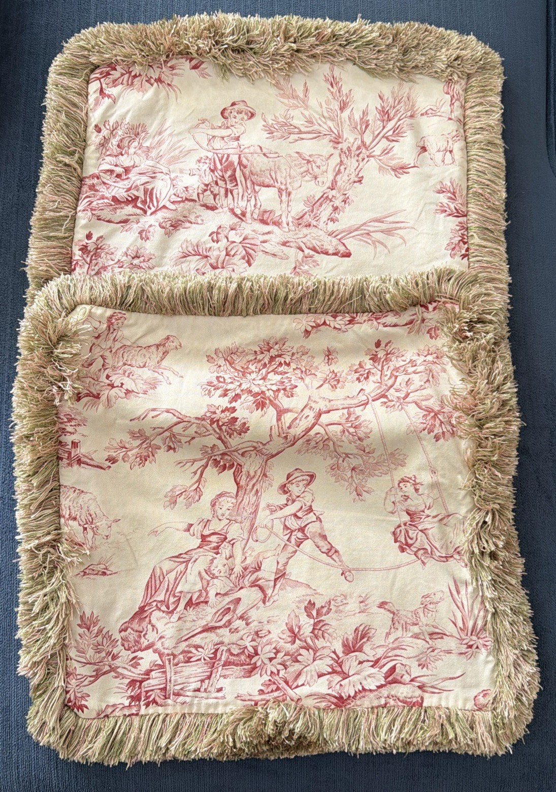 Two beige and red fringed toile pillow covers with pastoral print.