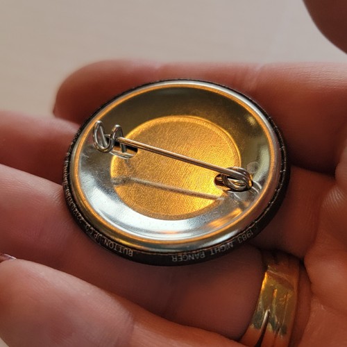 Round gold-tone pin back with black border and silver metal pin, made in USA, held in a person's hand.
