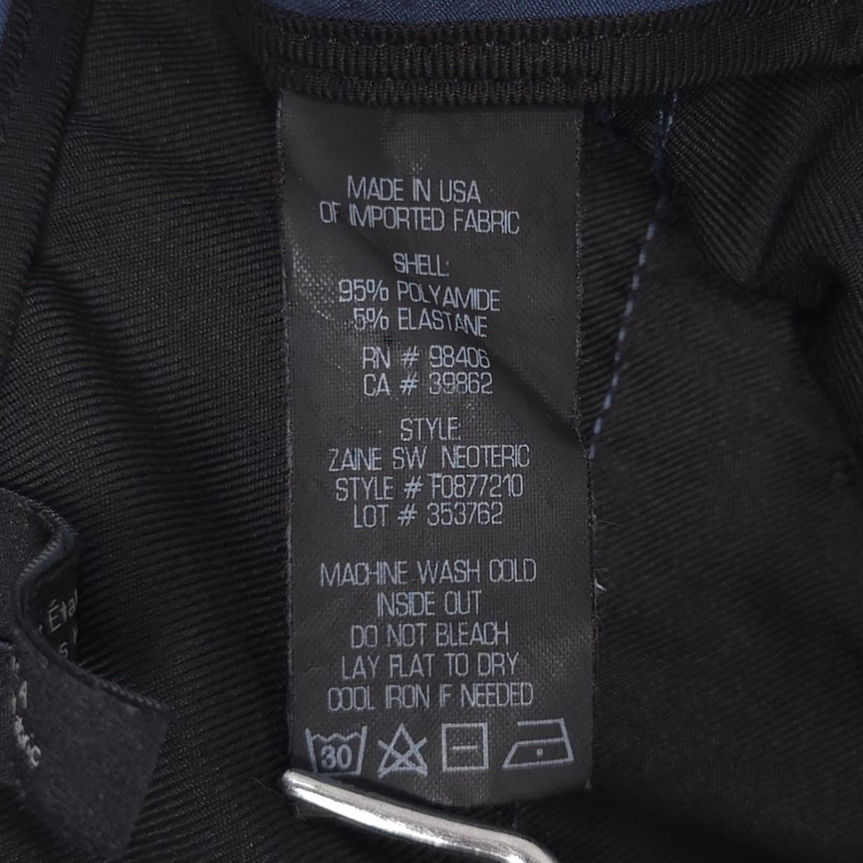 White clothing tag on black fabric with blue ZAINE SW NEOTERIC F0877210 label reading “Made in USA” and product details.