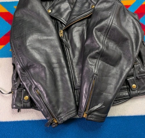 Black vintage leather Langlitz motorcycle jacket with front zipper, gold rivets, and visible stitching on a blue-red patterned surface.
