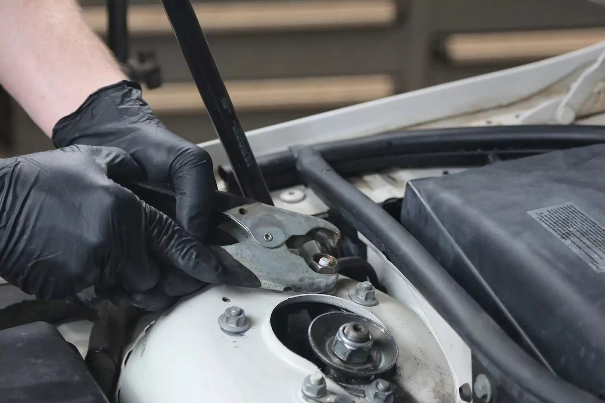 A mechanic wearing a black glove attaches a black handled jumper cable to a grounding point on a vehicle.
