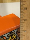 Orange Tupperware toy car with black roof, side graphic, and two yellow characters, measured against a wooden ruler.