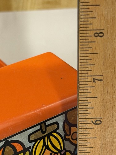 Orange Tupperware toy car with black roof, side graphic, and two yellow characters, measured against a wooden ruler.