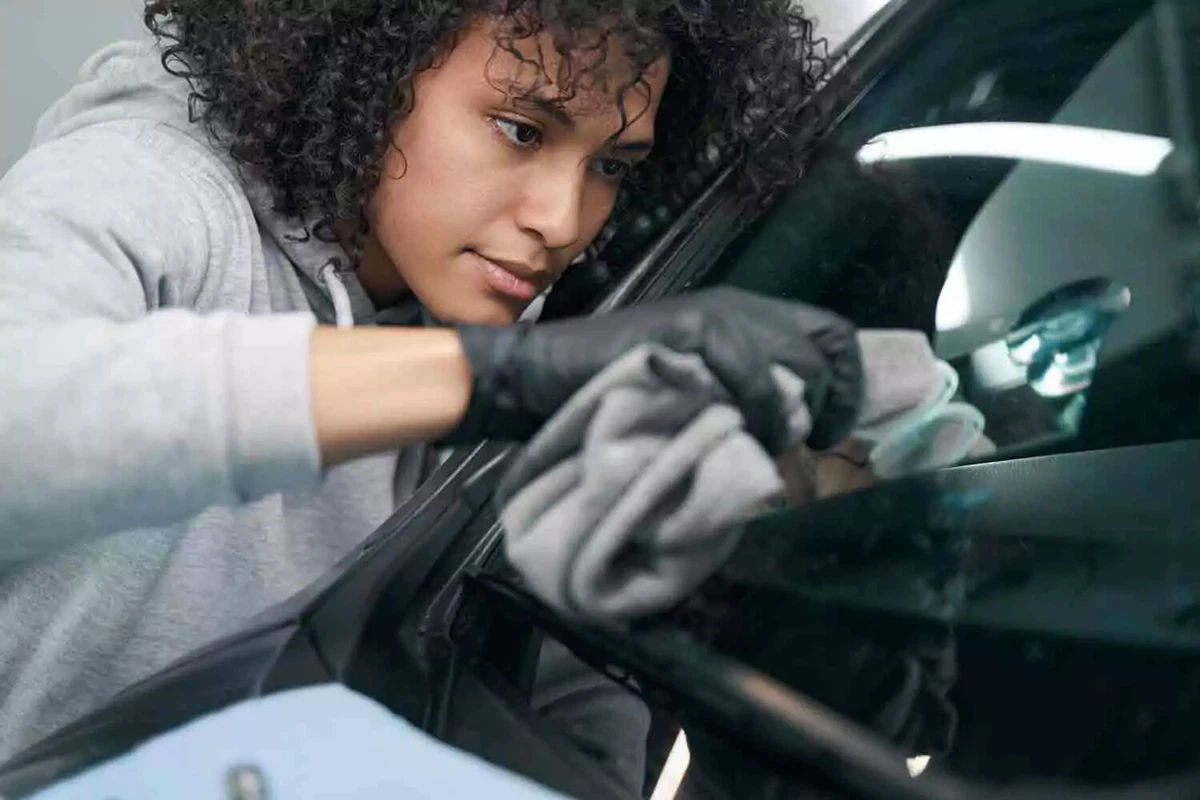 A technician wearing black nitrile gloves uses a gray microfiber towel to clean a windshield.