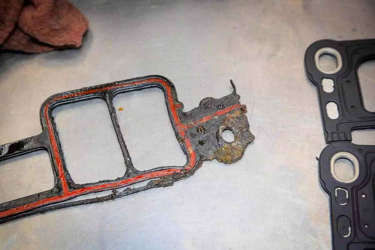 A close up view of an intake manifold gasket that has begun to fail, displayed on a metal work surface.