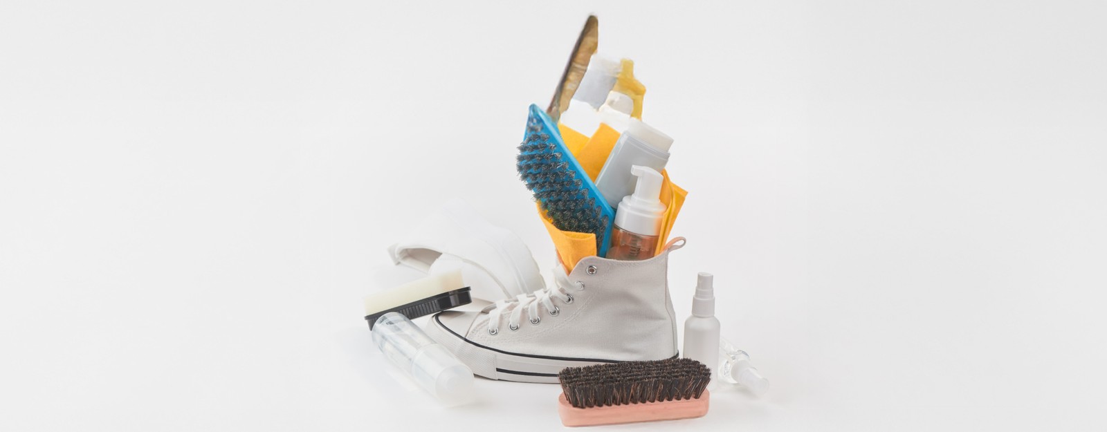 Vor einem weißen Hintergrund steht ein weiße Sneaker, umgeben von verschiedenen Pflegeutensilien wie Bürsten, Reinigungssprays, Tüchern und Flaschen. 