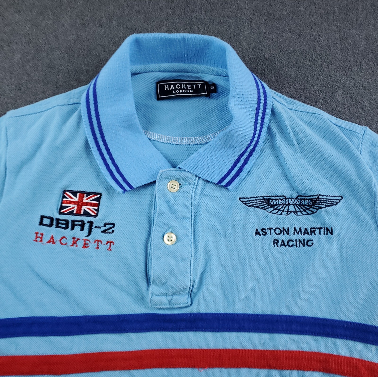 Light blue Hackett London polo shirt with navy trim, embroidered Aston Martin Racing logo, and Union Jack on chest.