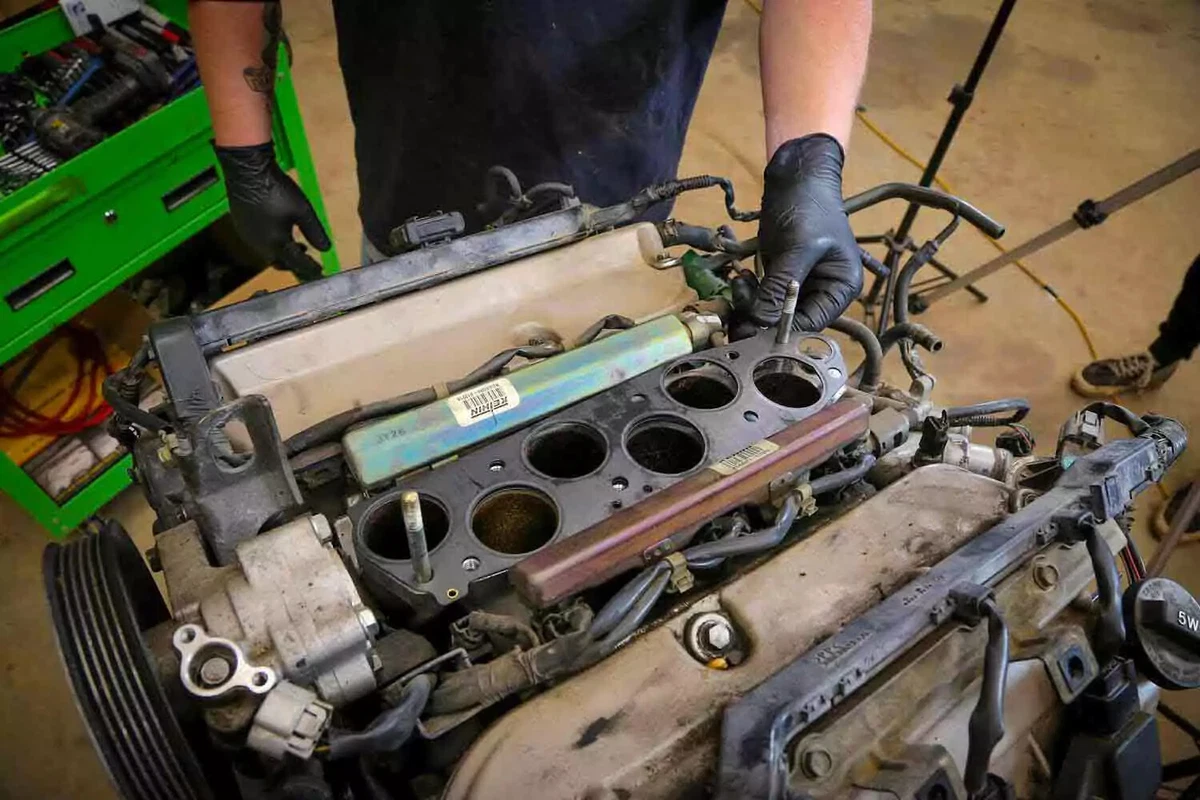 A mechanic wearing black nitrile gloves stands behind an early 2000s Honda V-6 engine, with the intake manifold and left-side cylinder head removed.