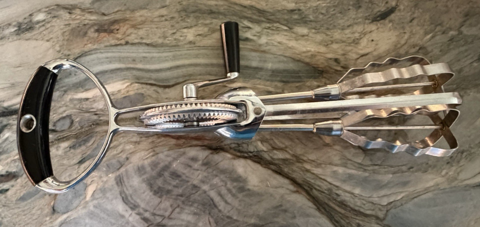 A Flint-brand manual egg beater with wavy blades and a sleek black handle — a visually striking piece of mid-century kitchen engineering that actually works and looks great on a hook.
