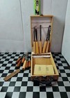 11 chisels with wooden handles in a brown wooden box, next to a yellow-edged oilstone and sharpening stone.