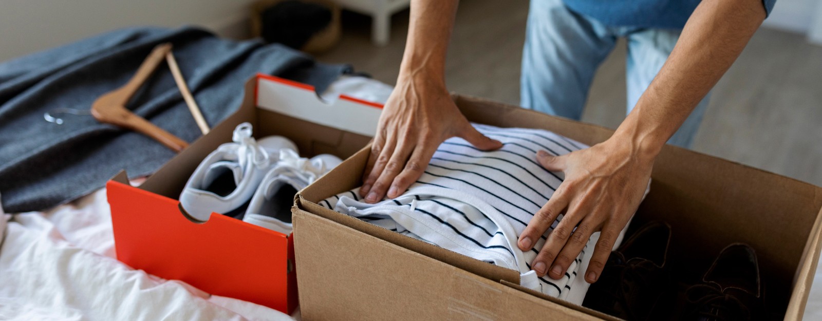 Eine Person packt zusammengelegte Kleidung und Schuhe in einen Karton auf einem Bett – eine Vorbereitung auf einen Umzug oder die Einlagerung.