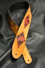 Brown leather guitar strap with red and black checkered inlay, featuring two silver rivets, on black fabric background.