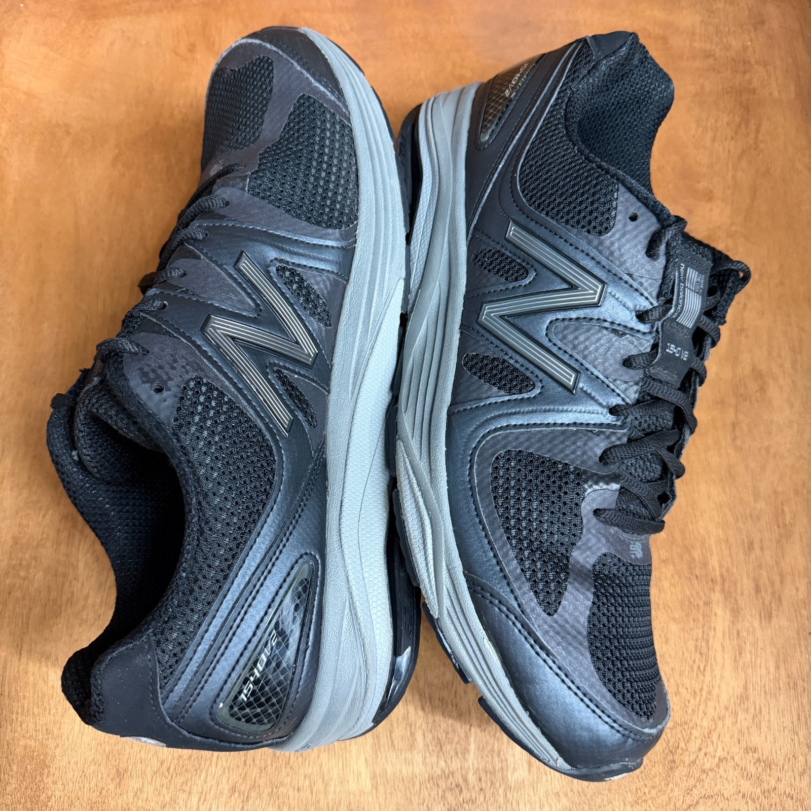 Black and gray New Balance 1540V2 men’s running shoes with silver “1540V2” text, mesh uppers, and gray laces on wooden floor.