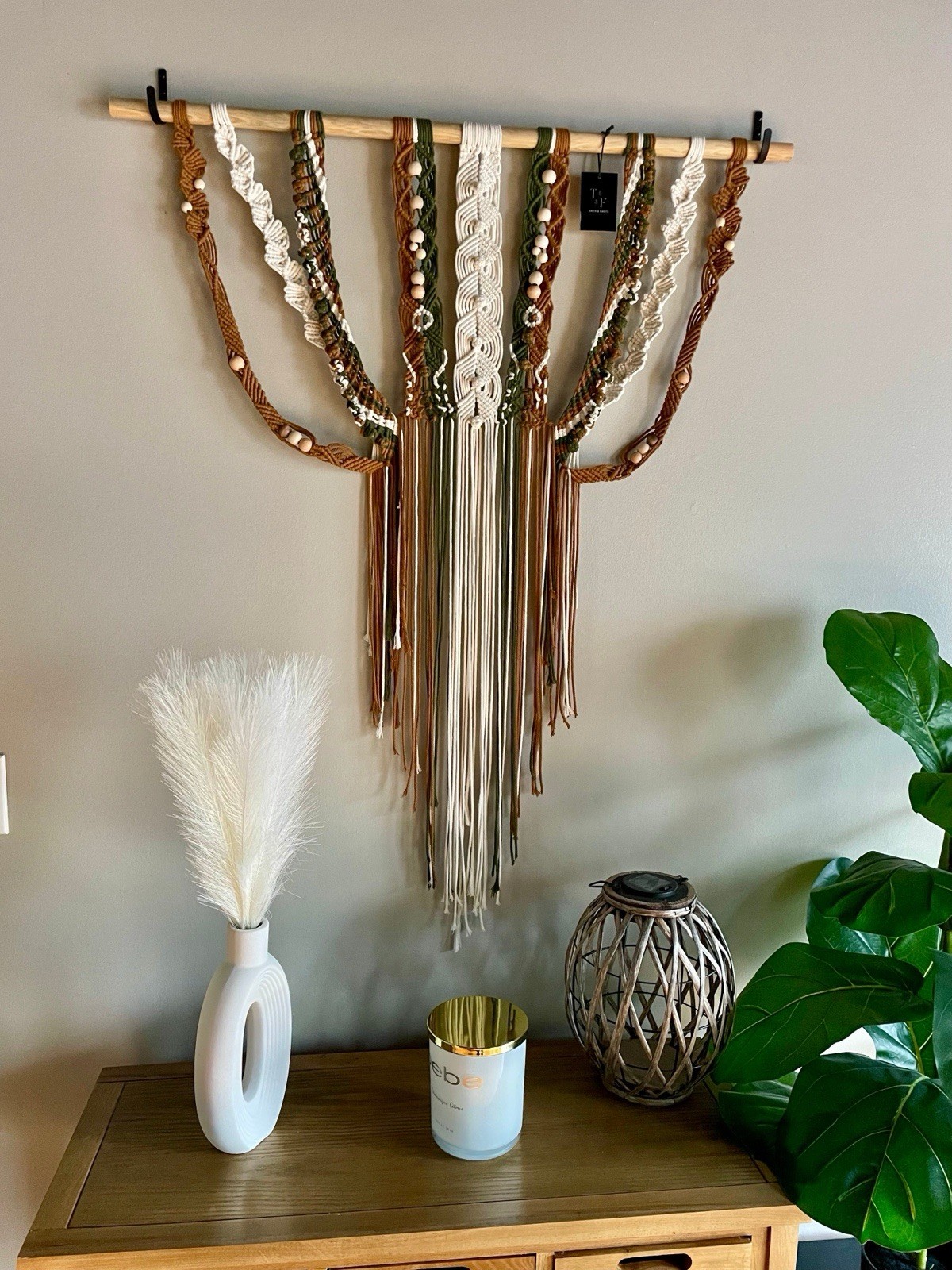 Brown, green, and cream boho macrame wall hanging with vertical stripes and fringe, displayed against a gray wall.