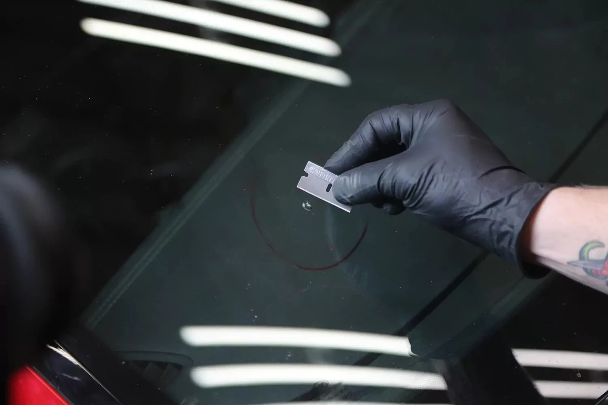 A mechanic wearing a black glove uses a razor blade to scrape away debris around a chip in a vehicle's windshield.
