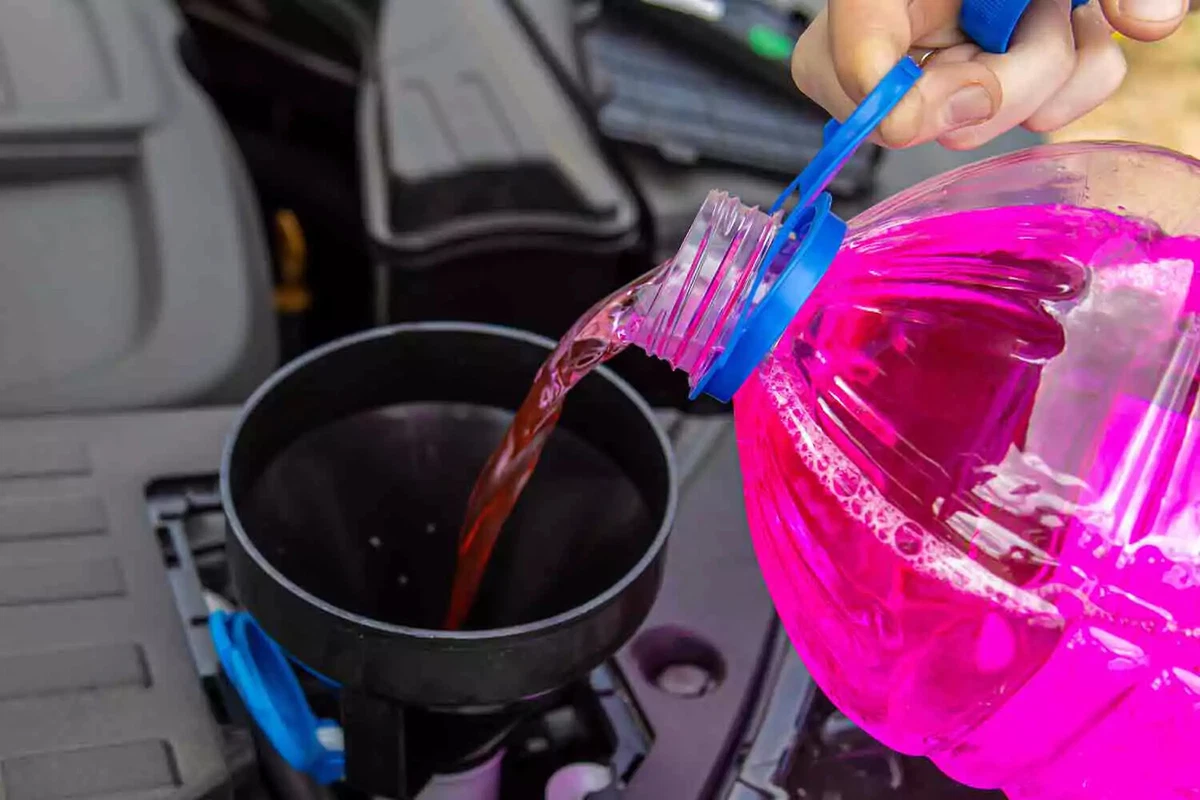 A driver pouring pink windshield washer fluid into a car’s windshield fluid reservoir through a black plastic funnel.