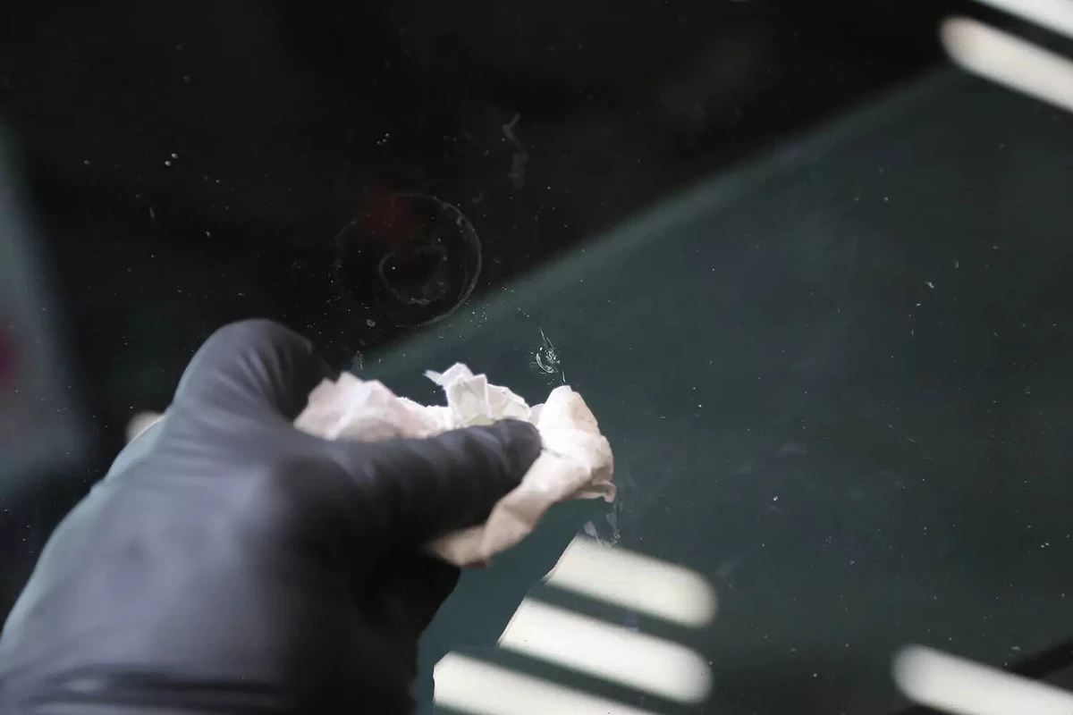 A mechanic wearing a black glove uses a white towel to wipe away excess repair liquid from a windshield.