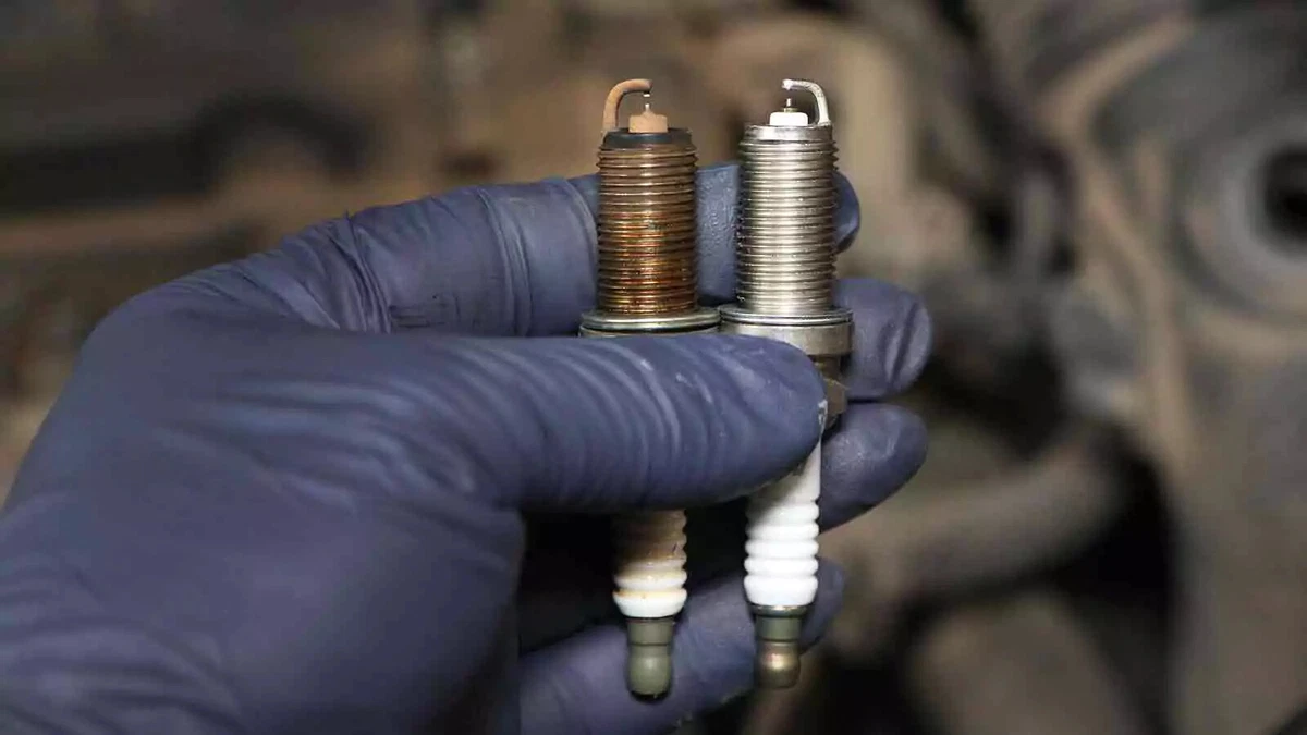 A mechanic wearing a blue nitrile glove holding two spark plugs, one used, one new.