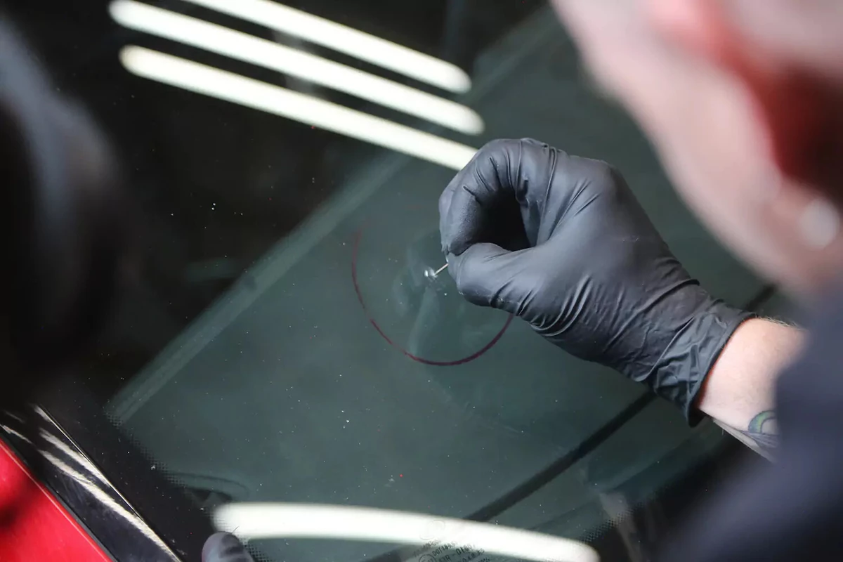 A mechanic wearing a black glove uses a push-pin tack to clean out bits of glass from a chip in a vehicle's windshield.