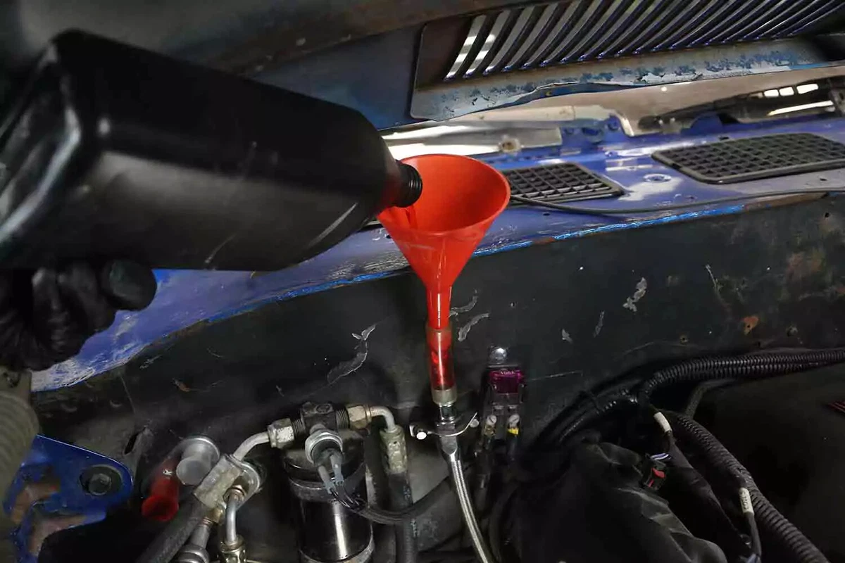 A mechanic wearing a black glove pours new transmission fluid into a red funnel to refill the vehicle's transmission.