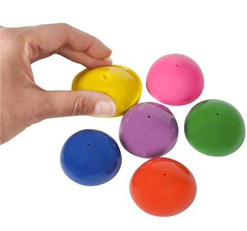Hand holding yellow plastic popper ball above five other multicolored popper balls on white background.