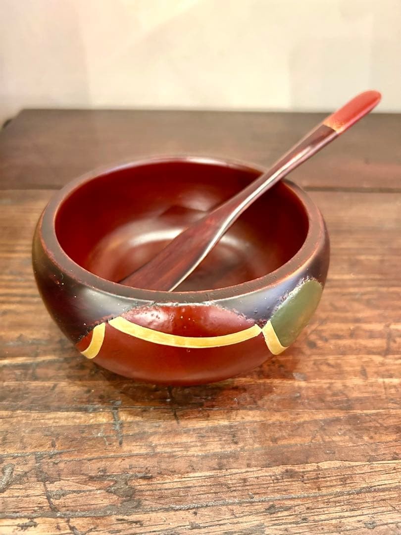 Red lacquerware bowl with brown rim, yellow band, and wooden spoon on rustic wooden surface.
