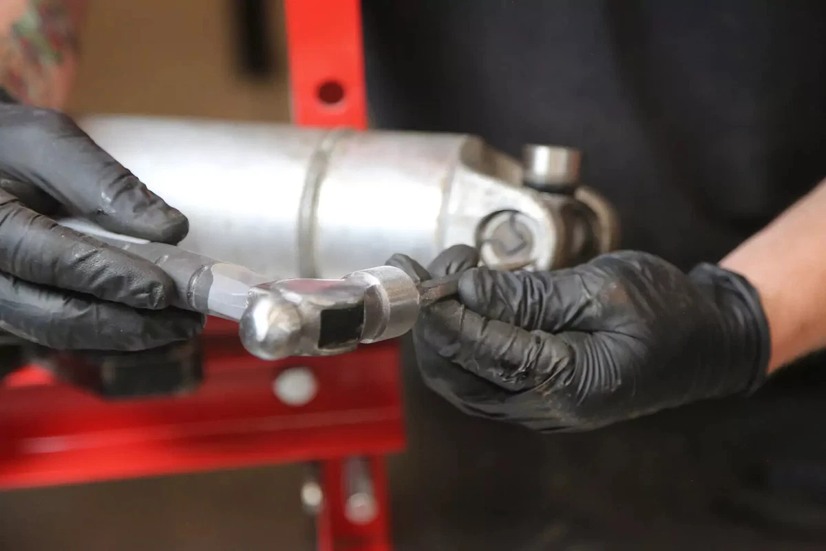 A mechanic wearing black gloves uses a small punch and hammer to set both clips into the grooves of a U-joint.