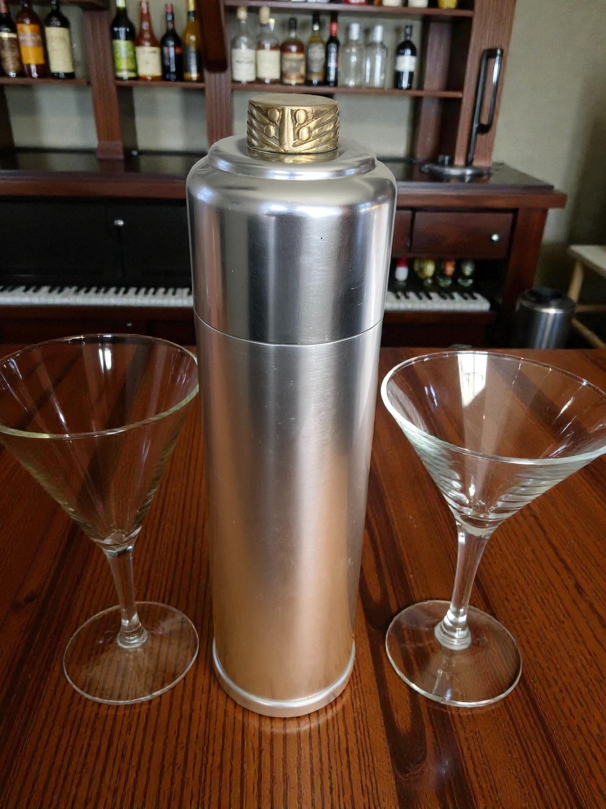 Brushed metal cocktail shaker with a ribbed gold-toned cap standing between two etched martini glasses on a wood bar counter.