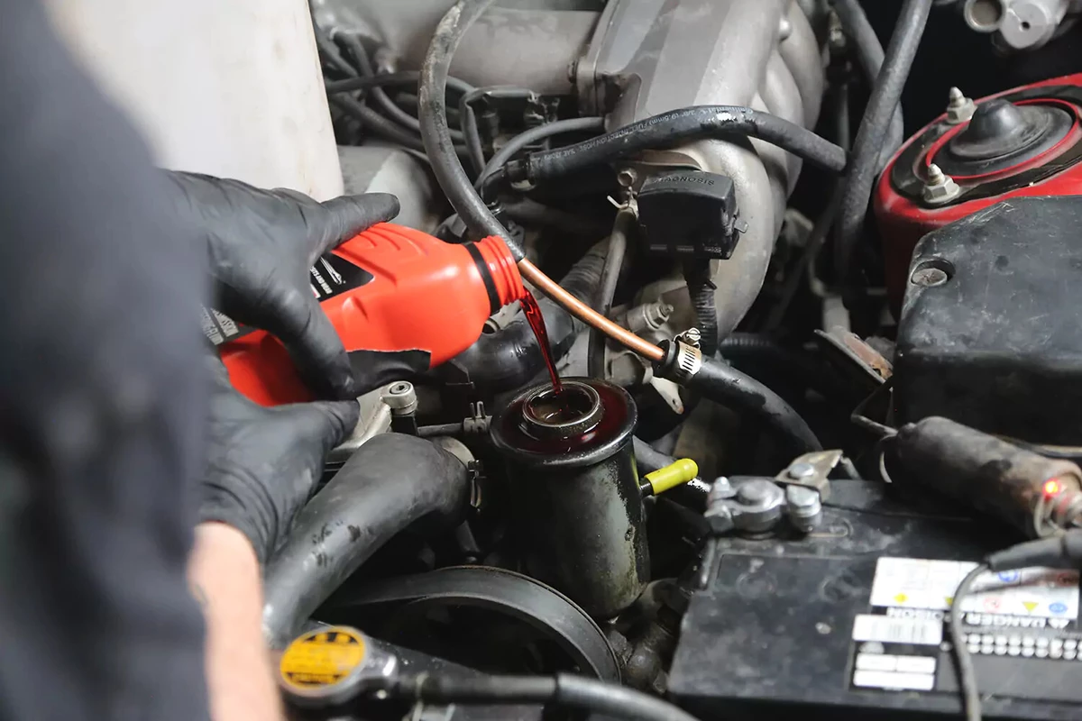 A mechanic wearing black gloves pours power steering fluid from a red bottle into a car's power steering fluid reservoir.